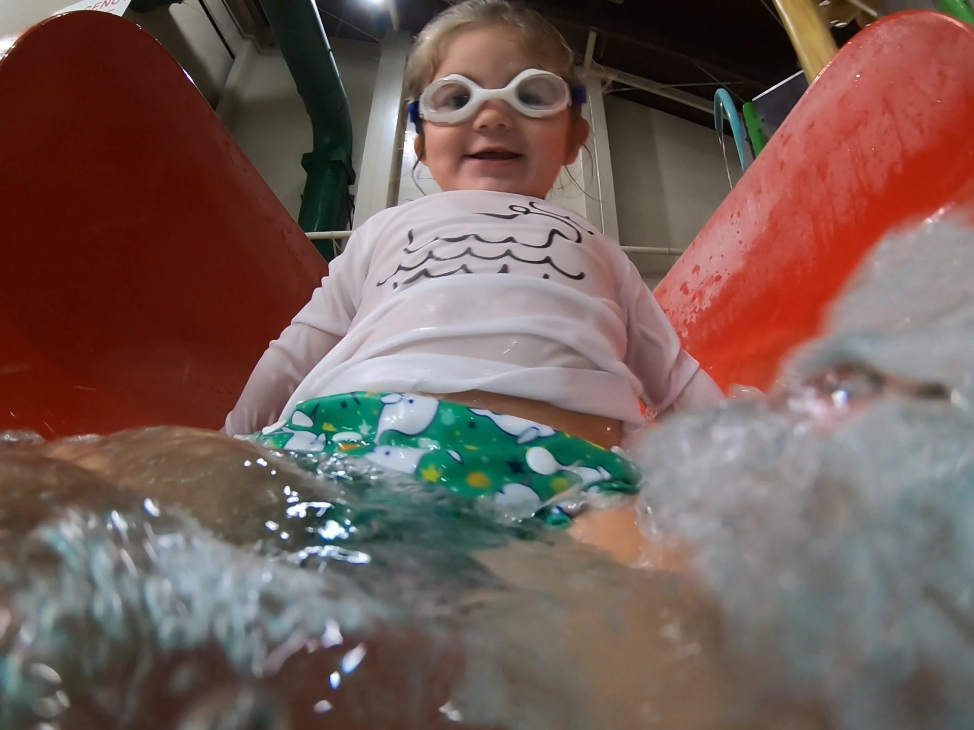 toddler girl at the bottom of a water slide wearing a Beau and Belle Littles Swim Diaper, Regular Size, green background, narwhals 