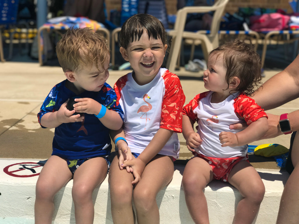three children sitting on the edge of the pool - two girls wearing the Beau and Belle Littles Rash Guard/Swim Shirt, white with pink 3/4 sleeves, flamingo print, one boy wearing  a Beau and Belle Littles Rash Guard/Swim Shirt, dark blue with lighter blue 3/4 sleeves, shark print