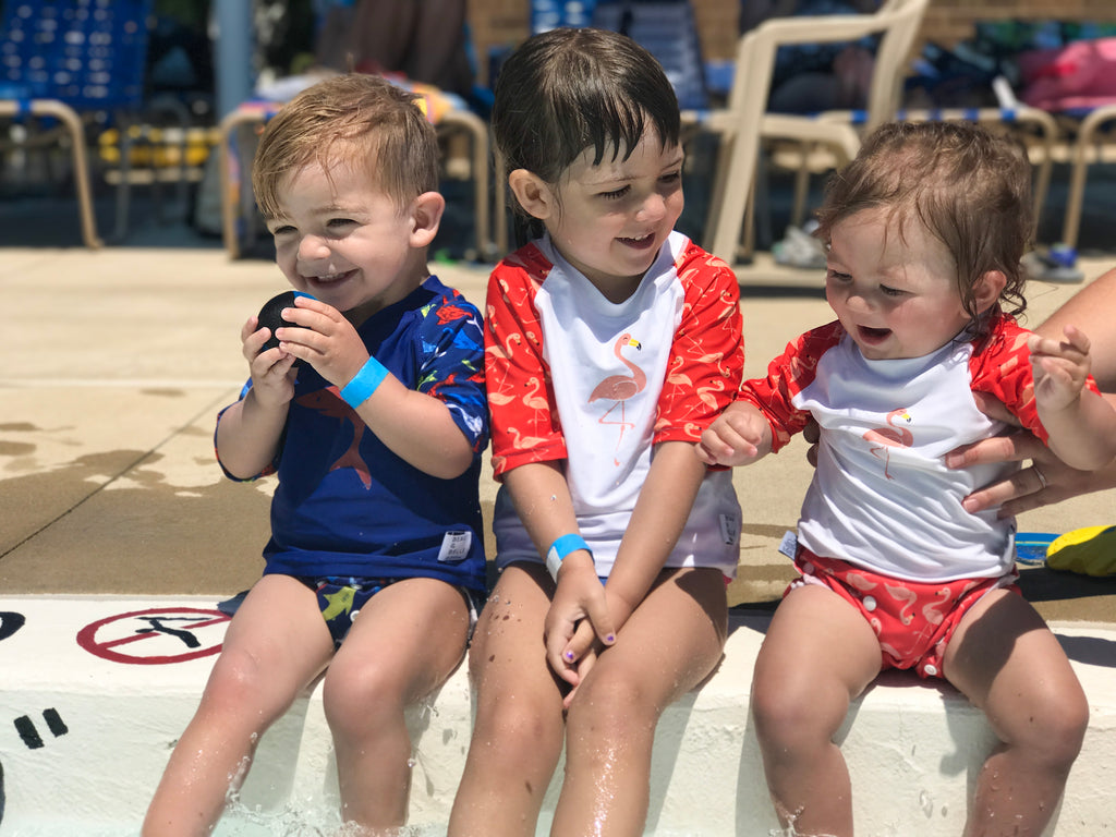 three children laughing and sitting on the edge of the pool - two girls wearing the Beau and Belle Littles Rash Guard/Swim Shirt, white with pink 3/4 sleeves, flamingo print, one boy wearing a Beau and Belle Littles Rash Guard/Swim Shirt, dark blue with lighter blue 3/4 sleeves, shark print
